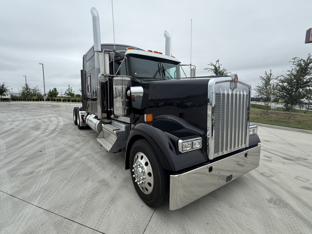 Kenworth W900L truck front-left view with shiny chrome grille and bumper. Large fuel tanks and dual tall exhaust stacks. No visible text on image aside from branding on the truck.