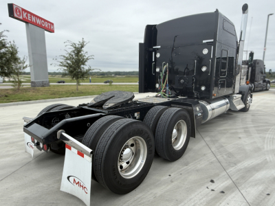 Kenworth W900L truck chassis rear view showing triple-axle configuration and fuel tanks, cab with sleeper, dealership sign in background.