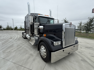 Kenworth W900L truck front-left view with shiny chrome grille and bumper. Large fuel tanks and dual tall exhaust stacks. No visible text on image aside from branding on the truck.