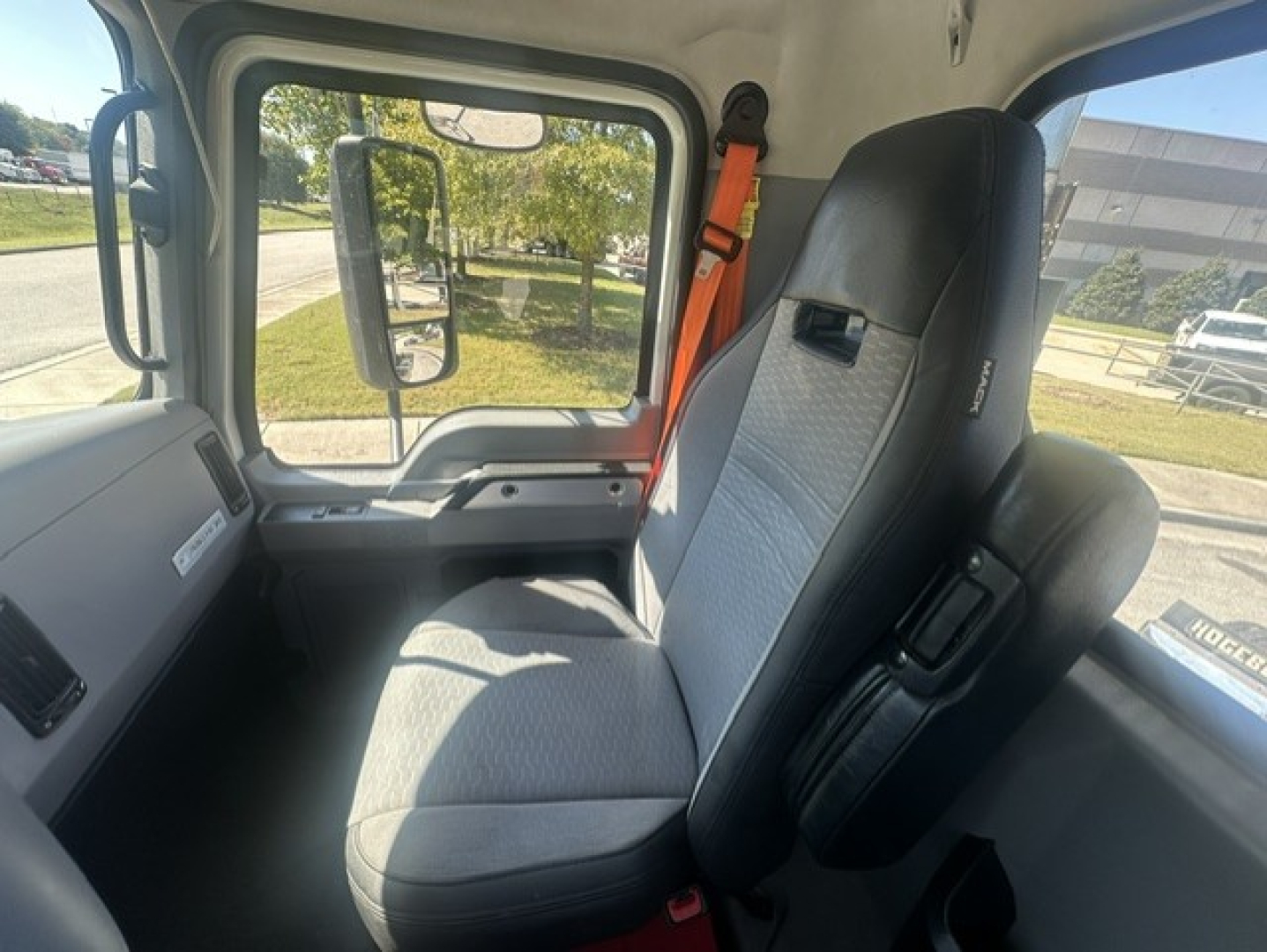 Interior of a Mack ANTHEM 64T truck cab showing driver seat and passenger seat with seat belts, dashboard window, and side mirror.