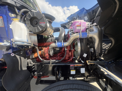 Mack PI64T engine bay showing coolant reservoir, pink coolant/DEF reservoir, red diesel engine, hoses, and yellow cap.