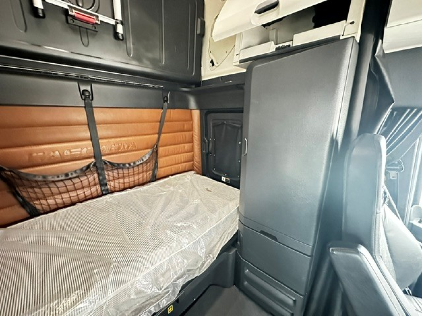 Freightliner Cascadia 126 interior sleeper cabin showing bunk bed with mattress wrapped in plastic, brown padded side panel, and black storage cabinets.