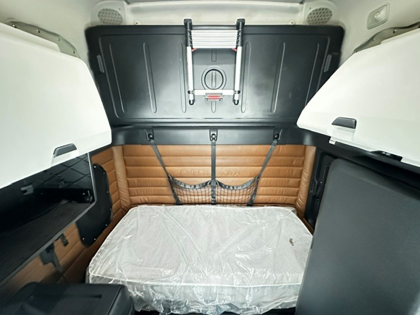 Truck cab interior showing sleeper area with brown padded wall, two storage compartments, and a plastic-covered mattress.