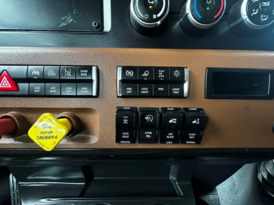 Cab dashboard panel of a Freightliner Cascadia showing various switches and knobs, including hazard button and yellow ignition start/stop button.