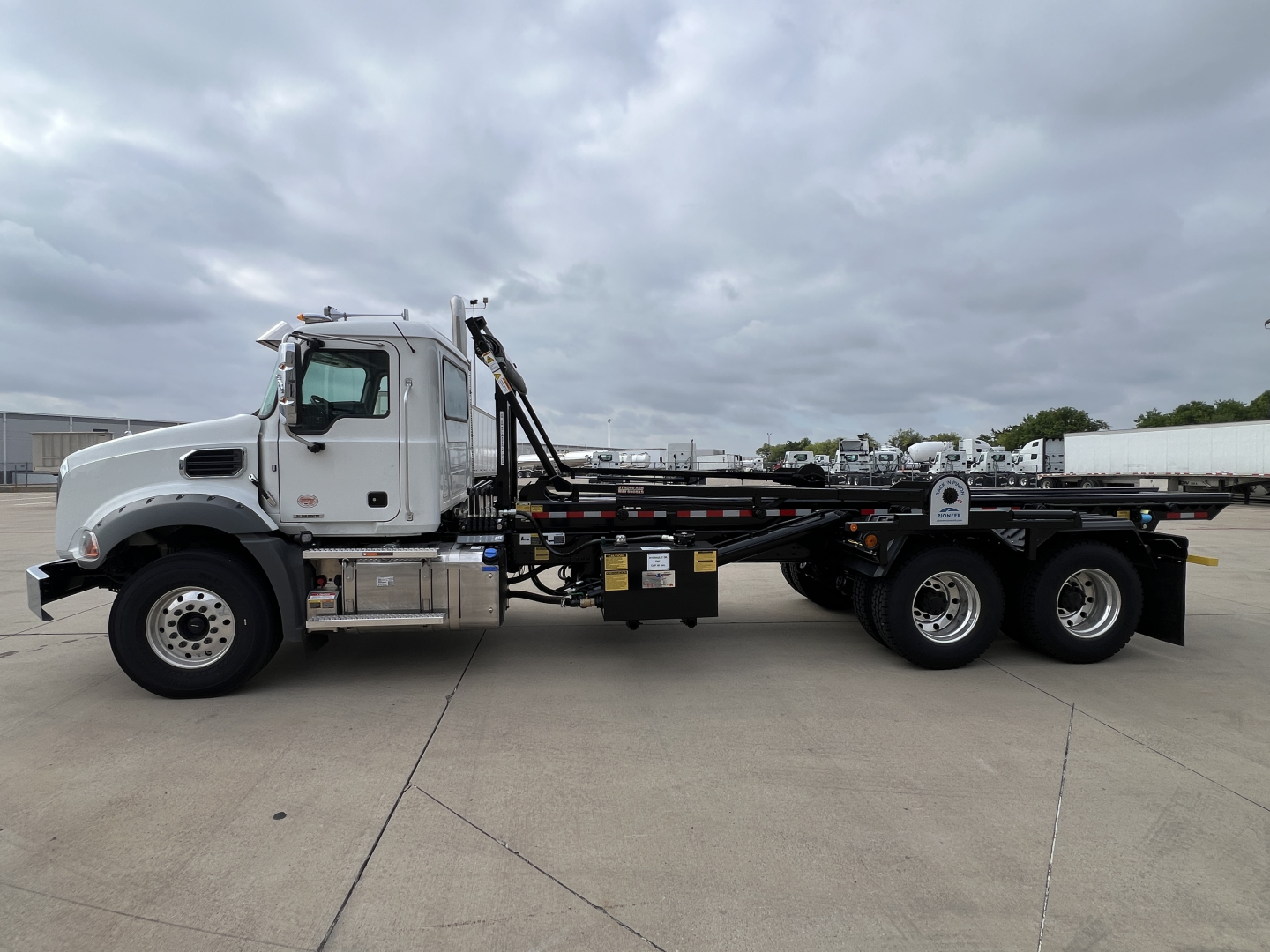 Side view of a new 2027 Mack GR64B roll-off truck showing chassis and triple rear axles with hydraulic roll-off lift system.