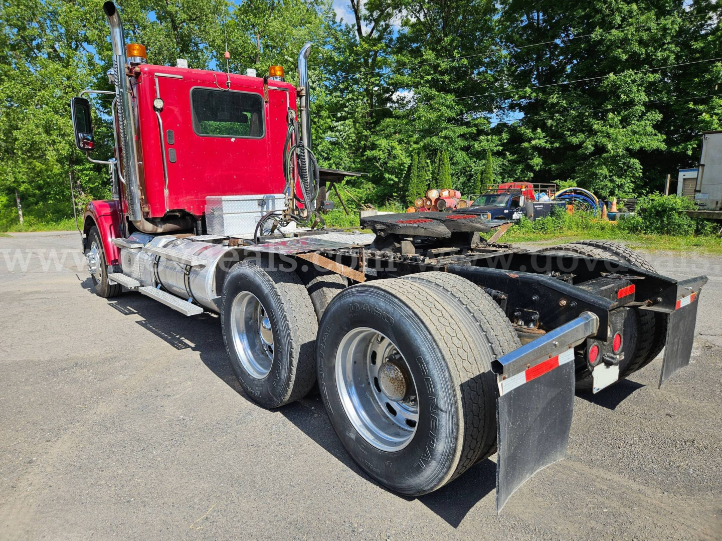 Red Freightliner Coronado SD semi-truck cab and chassis in a parking lot, rear view showing dual rear axles, fifth-wheel area, fuel tanks, and mud flaps.