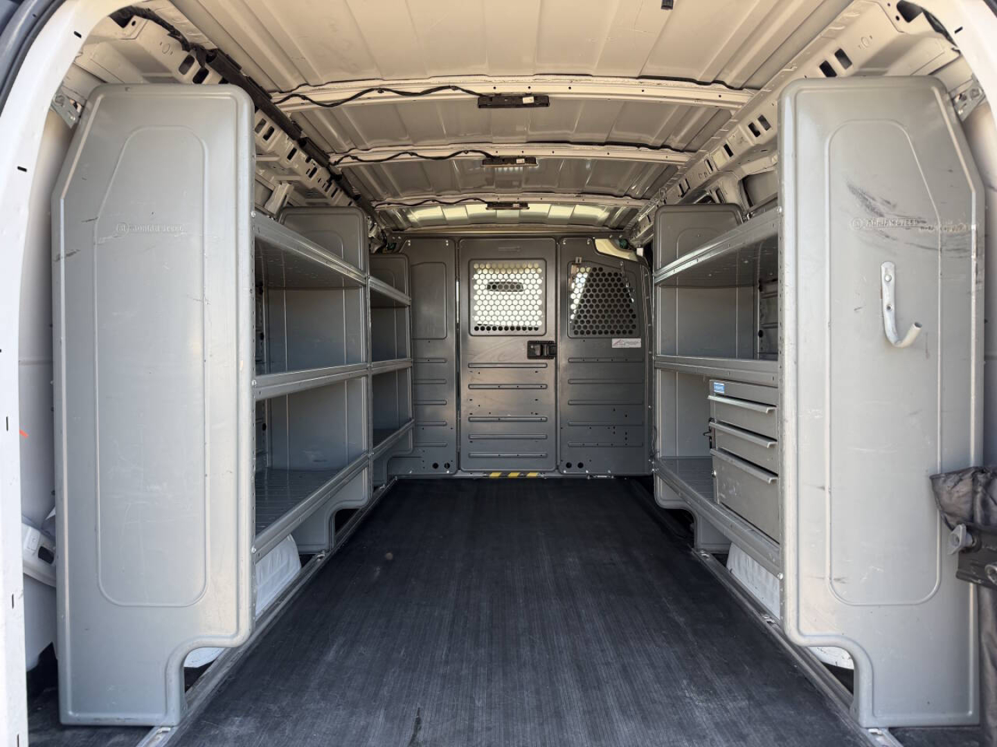 Interior of a 2022 Chevrolet Express cargo van showing empty shelving units along both sides, partition with metal panels, and a black rubber floor.