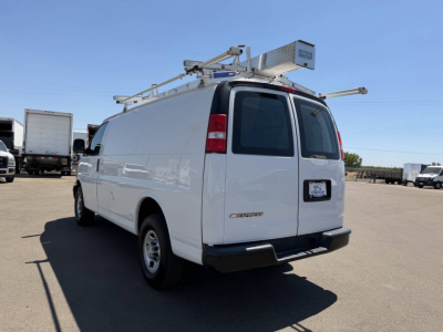 Chevrolet Express cargo van rear/side view with roof ladder rack and extension arms, white exterior, two back doors, blank rear license area.