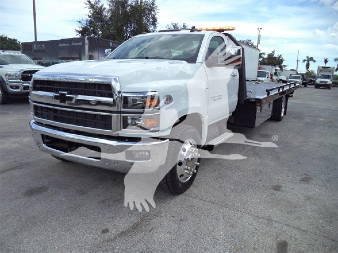 Chevrolet Silverado 6500HD tow truck with flatbed viewed from front-left, chrome grille, white cab, long wheelbase, daytime dealership lot.