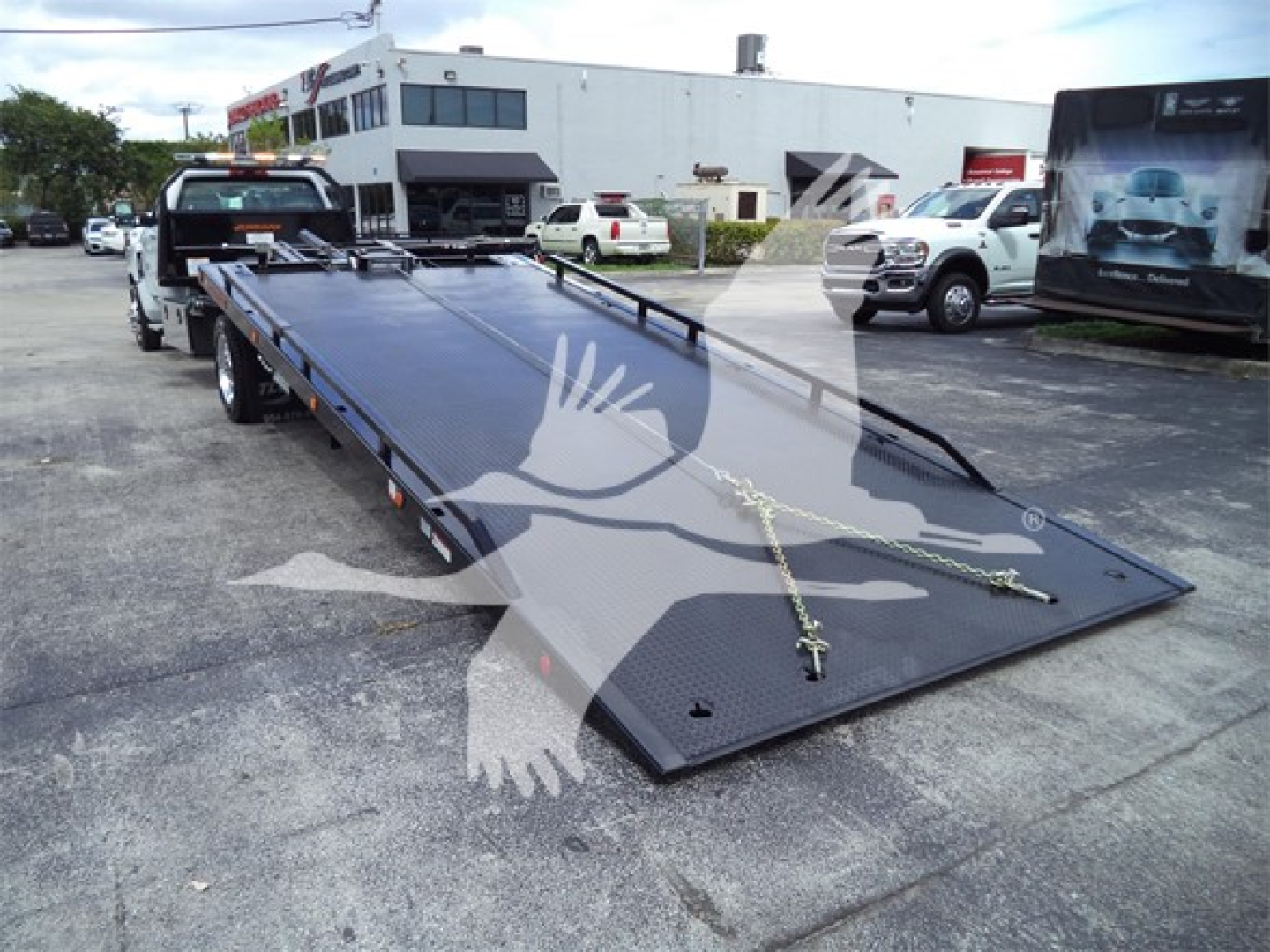 Truck rollback flatbed on a dual-axle trailer with a blue-black metal surface and chains, dealership lot in background.