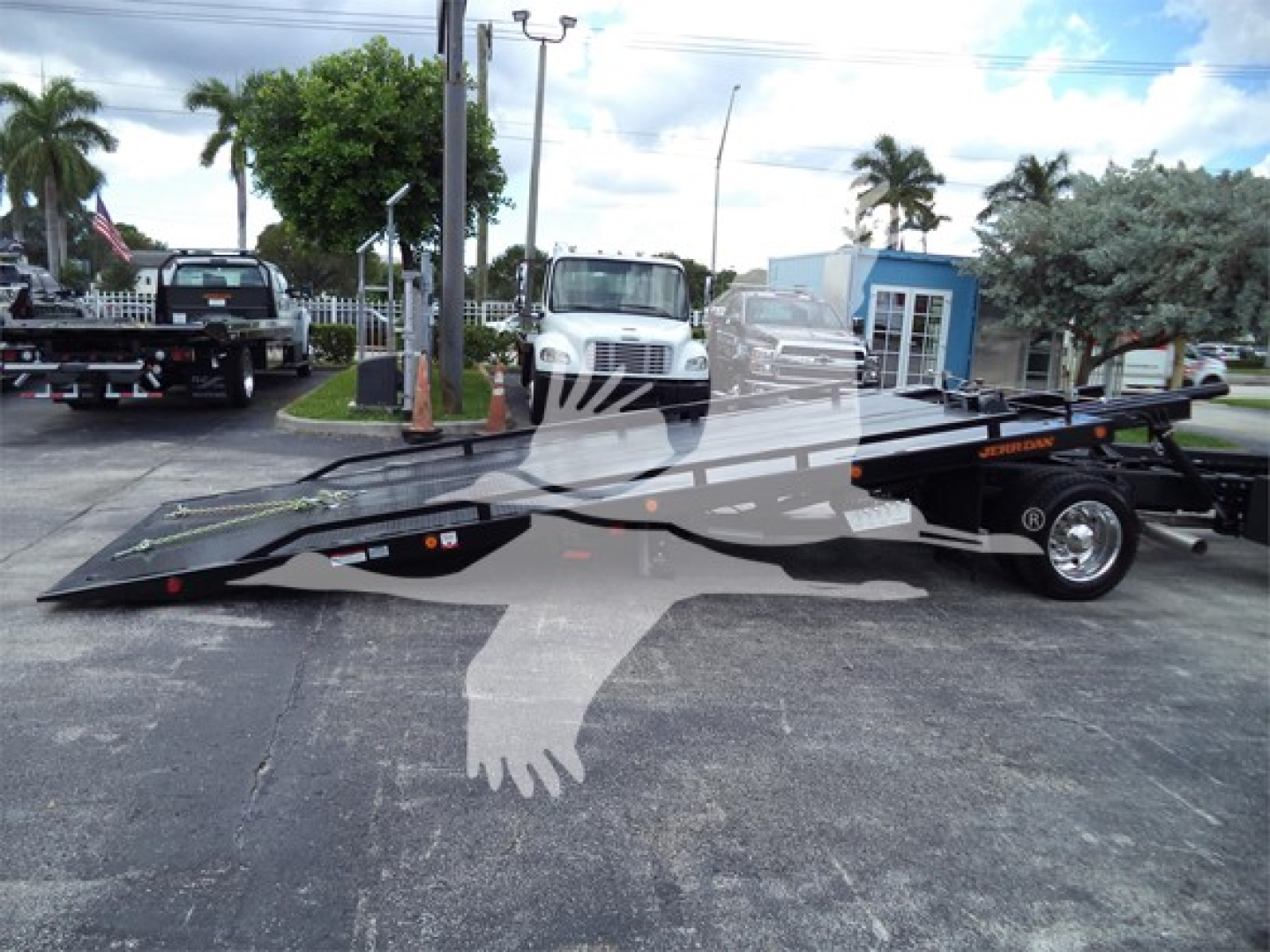 Truck flatbed tow truck in lot with Chevrolet Silverado 6500HD in background showing long steel tilt-bed and wheel chocks.