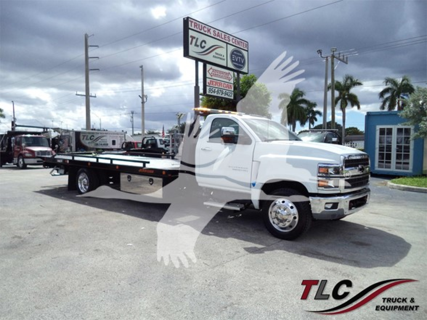Chevrolet Silverado 6500HD rollback tow truck in a dealership lot, white cab with flatbed, TLC Truck Sales Center signage in background.