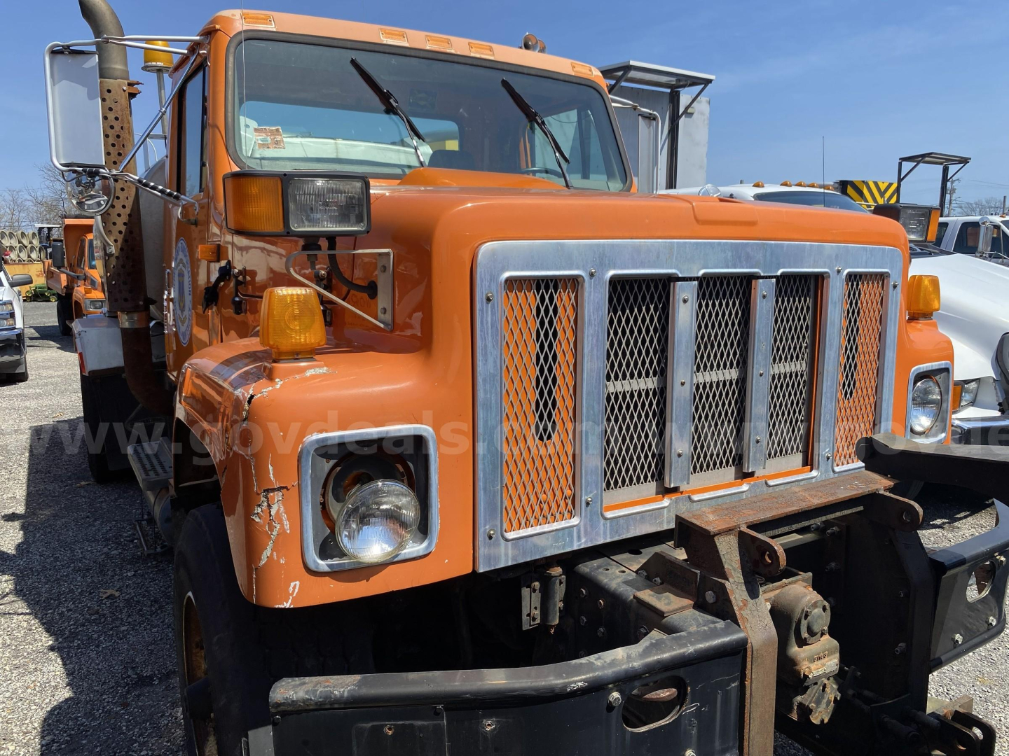 Orange 2002 International 2574 truck front view showing grille, bumper, headlights, and cab; visible metal radiator guard and side mirror; multiple utility features.