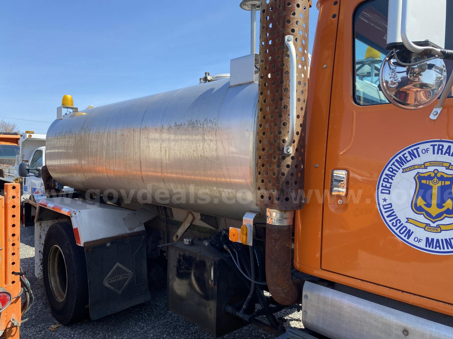 Tank truck with a large cylindrical stainless steel tank mounted on a chassis, orange cab door visible with Department of Transportation logo, reflection on tank.