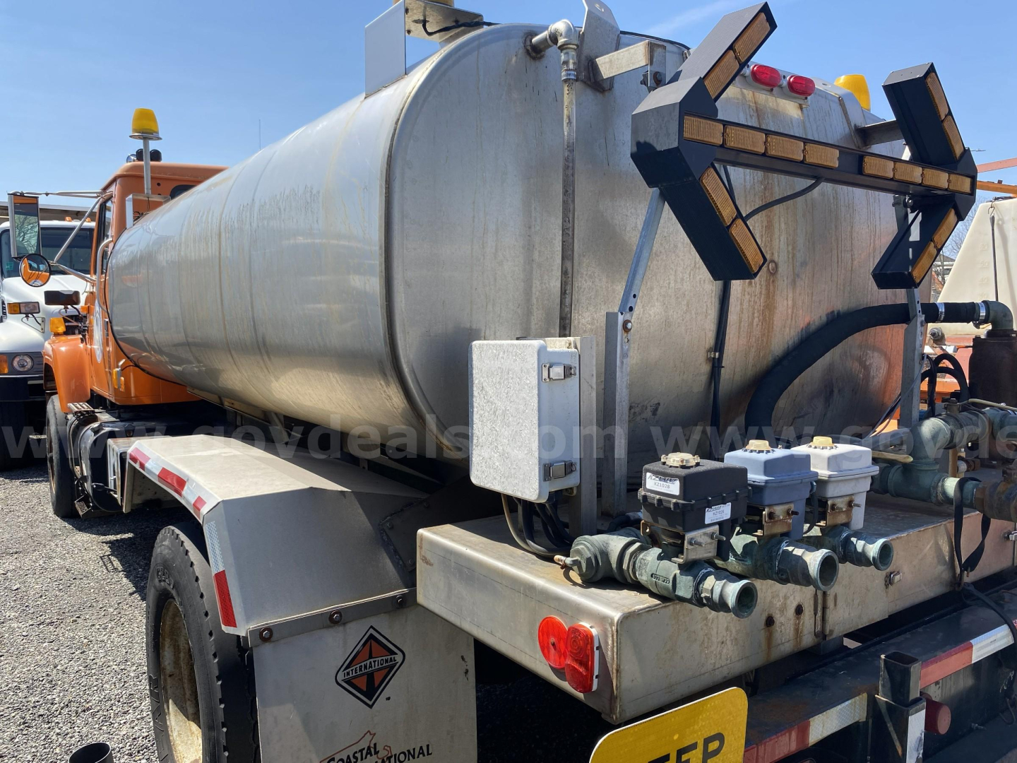 Tank truck rear with cylindrical stainless steel tanker and valve controls. Platform shows warning label, lights, and piping assemblies.
