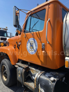 Truck cab side view showing orange International 2574 with emblem on door, fuel tank, and steps visible.