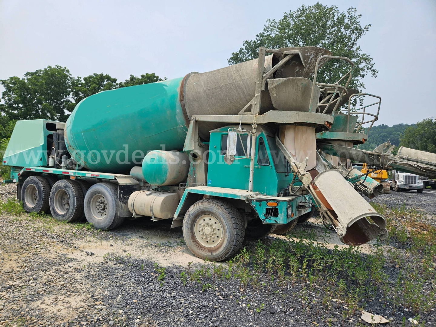 Mixer truck from 1999 Oshkosh S2346 with green cement mixer body and attached chute, visible dual-axle rear drive, worn outer body, and dirt road setting.