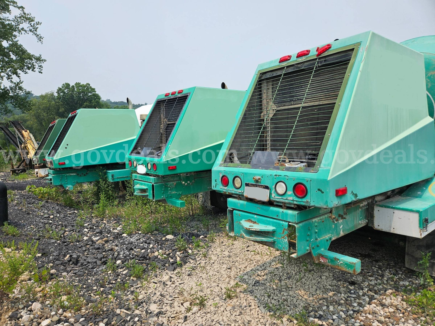 Three teal Oshkosh S2346 mixer trucks seen from rear-left side; visible vents/grilles and taillights; weathered equipment in a gravel lot.