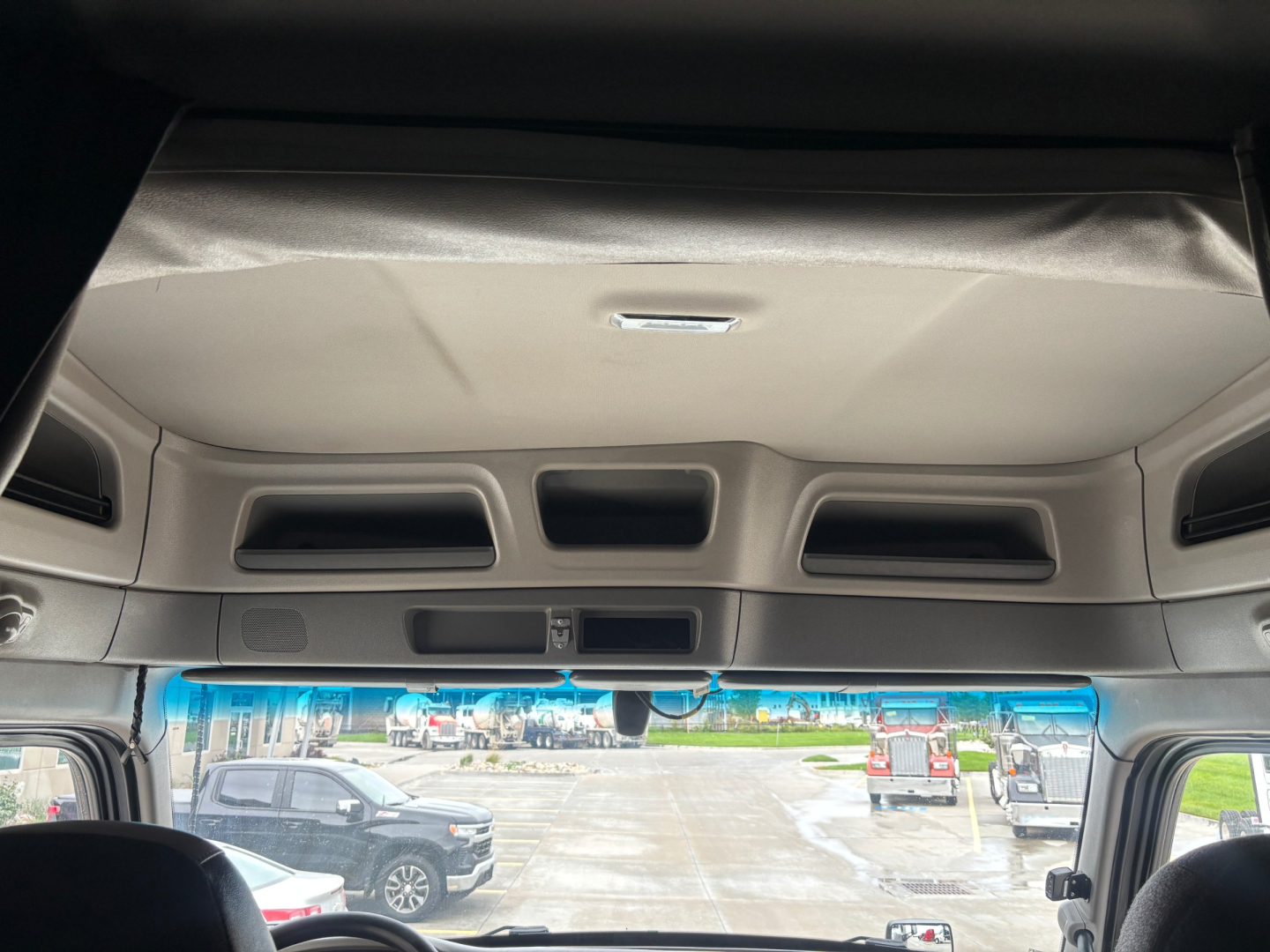 Truck cab ceiling and upper dash area of a Peterbilt 579 interior with overhead storage and sun visors; windshield view shows parking lot and other vehicles.