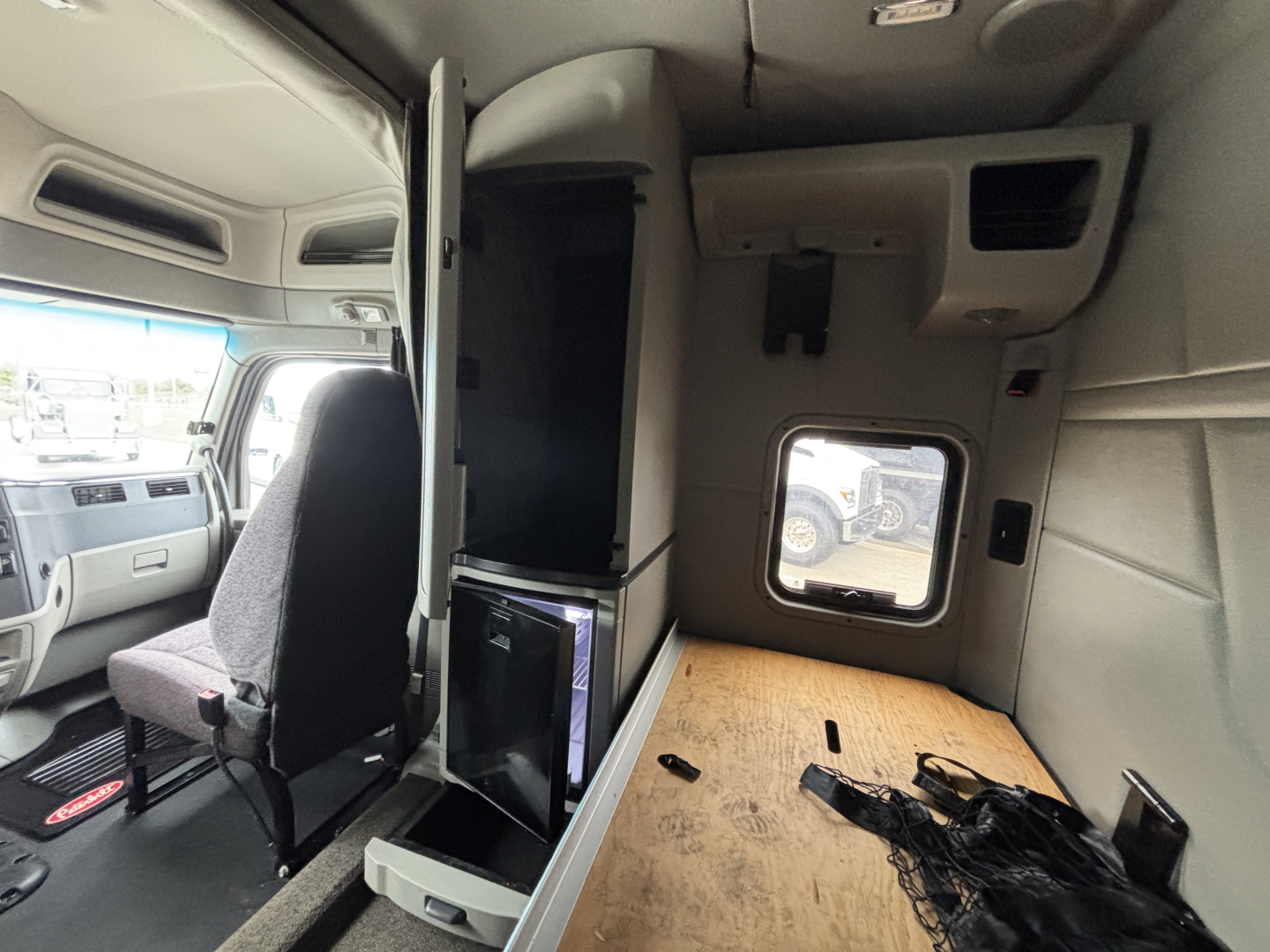 Peterbilt 579 cab interior showing sleeper area and upper storage with a small refrigerator, plywood floor panel, and cabin fixtures.