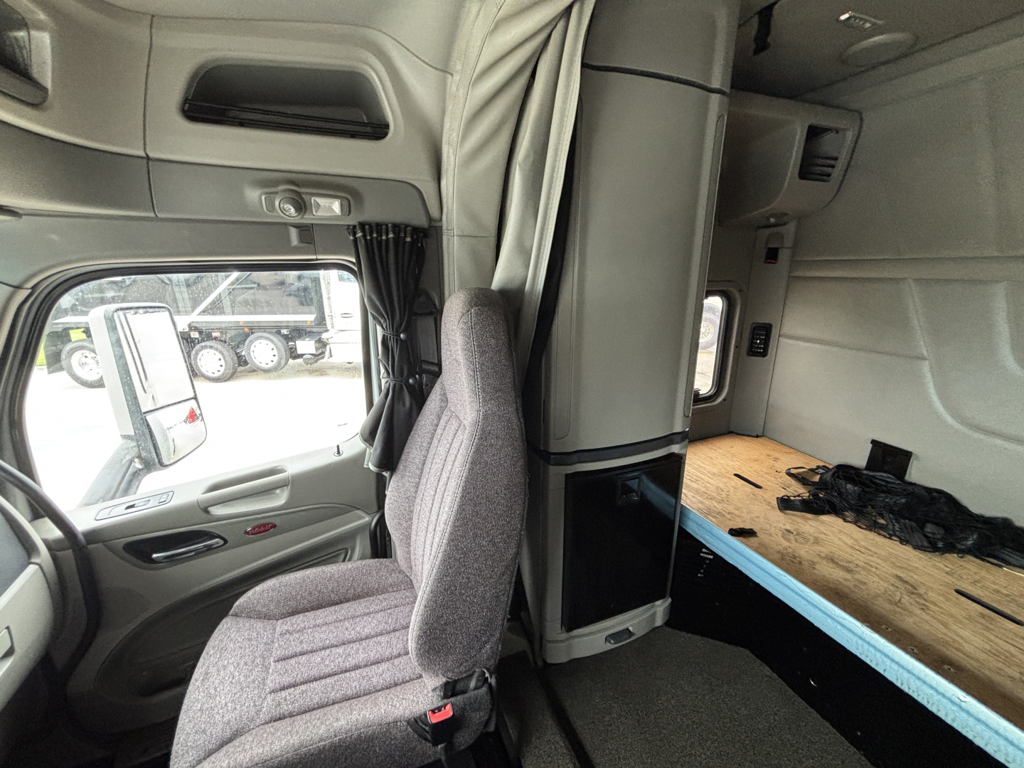 Truck cab interior of a Peterbilt 579 sleeper showing driver seat, window, and a wooden sleeper platform with tools.