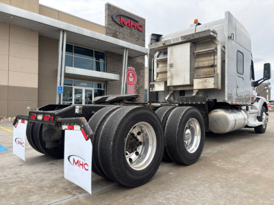 Peterbilt 579 tractor rear view showing dual rear axles and exhaust stack, with MHC dealership in background.