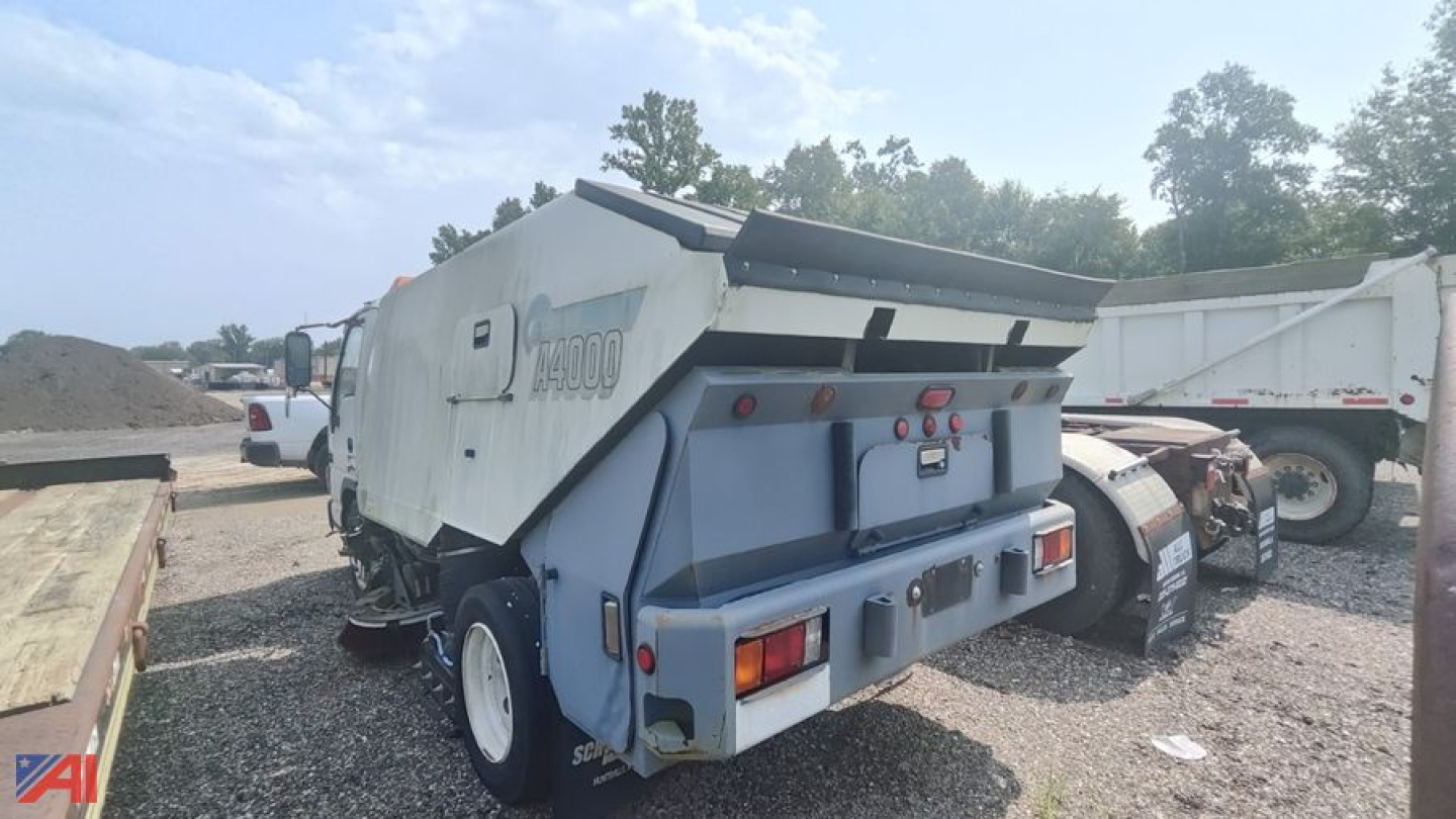 Rear-left view of a white/blue GMC Isuzu W5500 sweeper truck with hopper open, showing model label and rear lights.