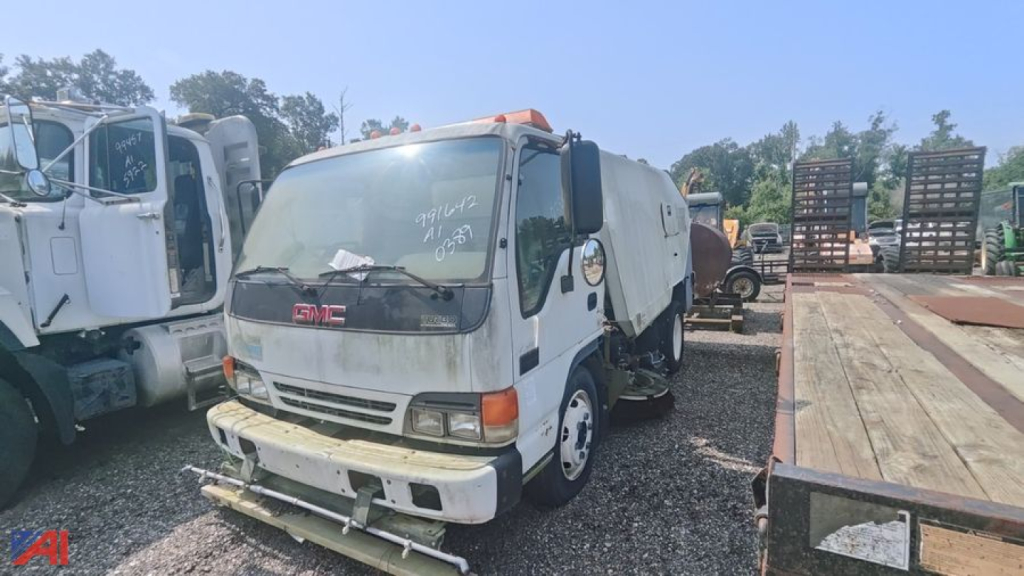 GMC Isuzu W5500 sweeper truck in a used lot, front view with white cab, swept body, orange beacon, and adjacent equipment on gravel.