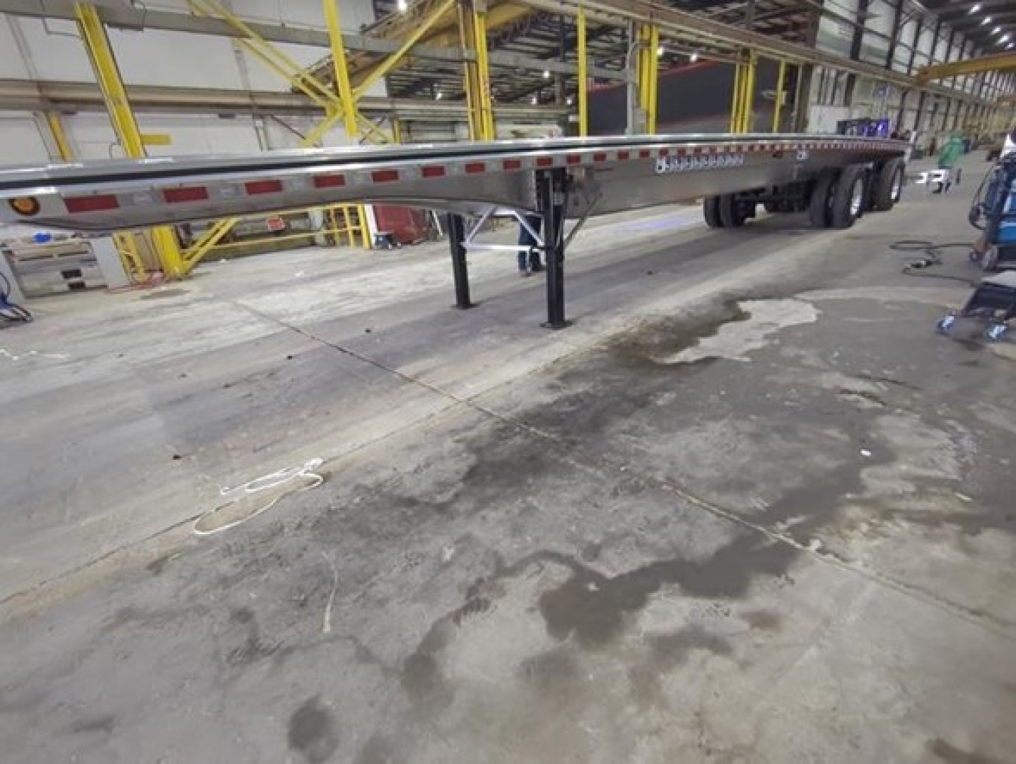 Trailer undercarriage showing flatbed trailer with support legs in factory setting; long silver frame with reflective markers