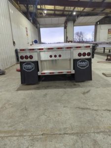 Mac trailer back end view showing multiple red marker lights, metal frame, mud flaps with MAC trailer logos, and reflective tape.