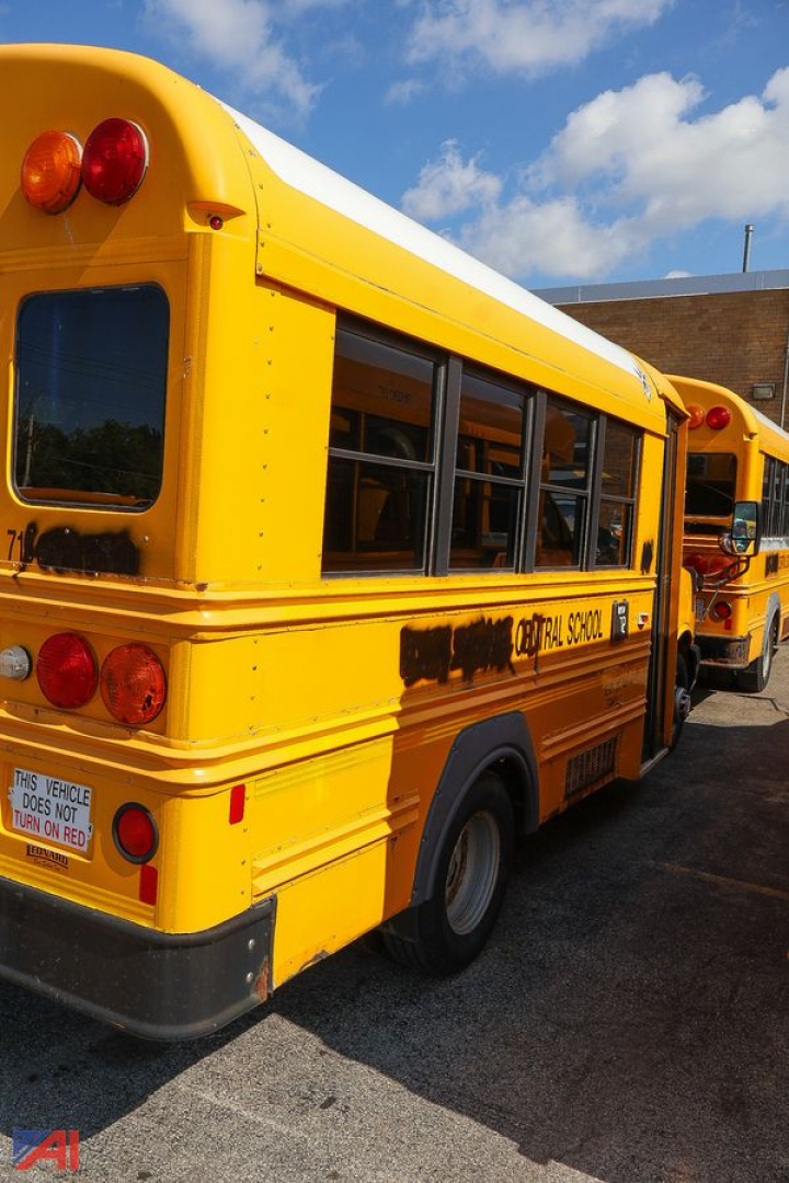 Rear and side view of a yellow school bus with black window frames, showing luggage and rear lights; text on side reads 'SCHOOL' with partial branding; blue sky with clouds in background.