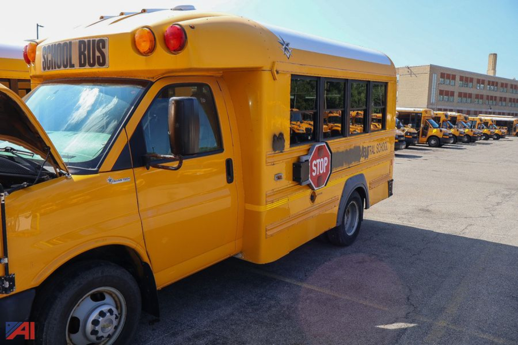 School bus exterior with open hood and 'SCHOOL BUS' sign on the roof, yellow Express/Trans Tech vehicle with stop sign and multiple similar buses in background.