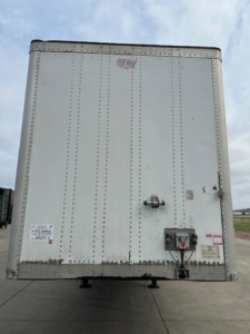 Rear view of a white cargo trailer with riveted panels showing the back doors, latch, and electrical connection; small labels and stickers on lower left and right.