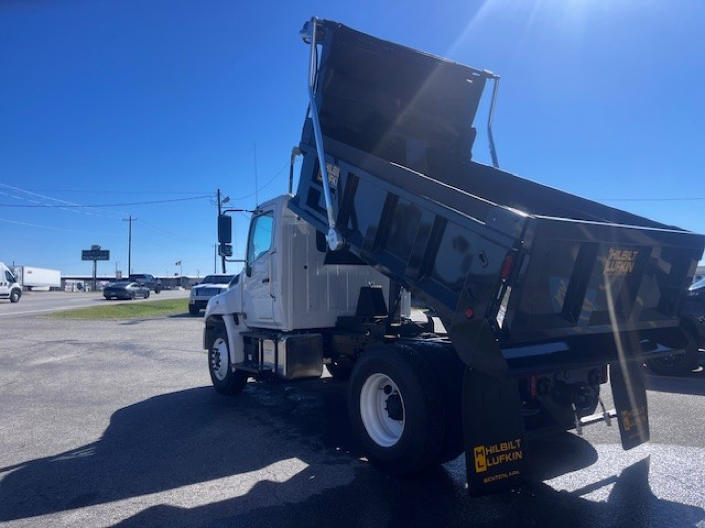 Truck with dump bed raised showing side view of cab and hydraulic dump mechanism. DL: white cab, black dump bed, rear dual tires, Hino branding not visible, clear sky.