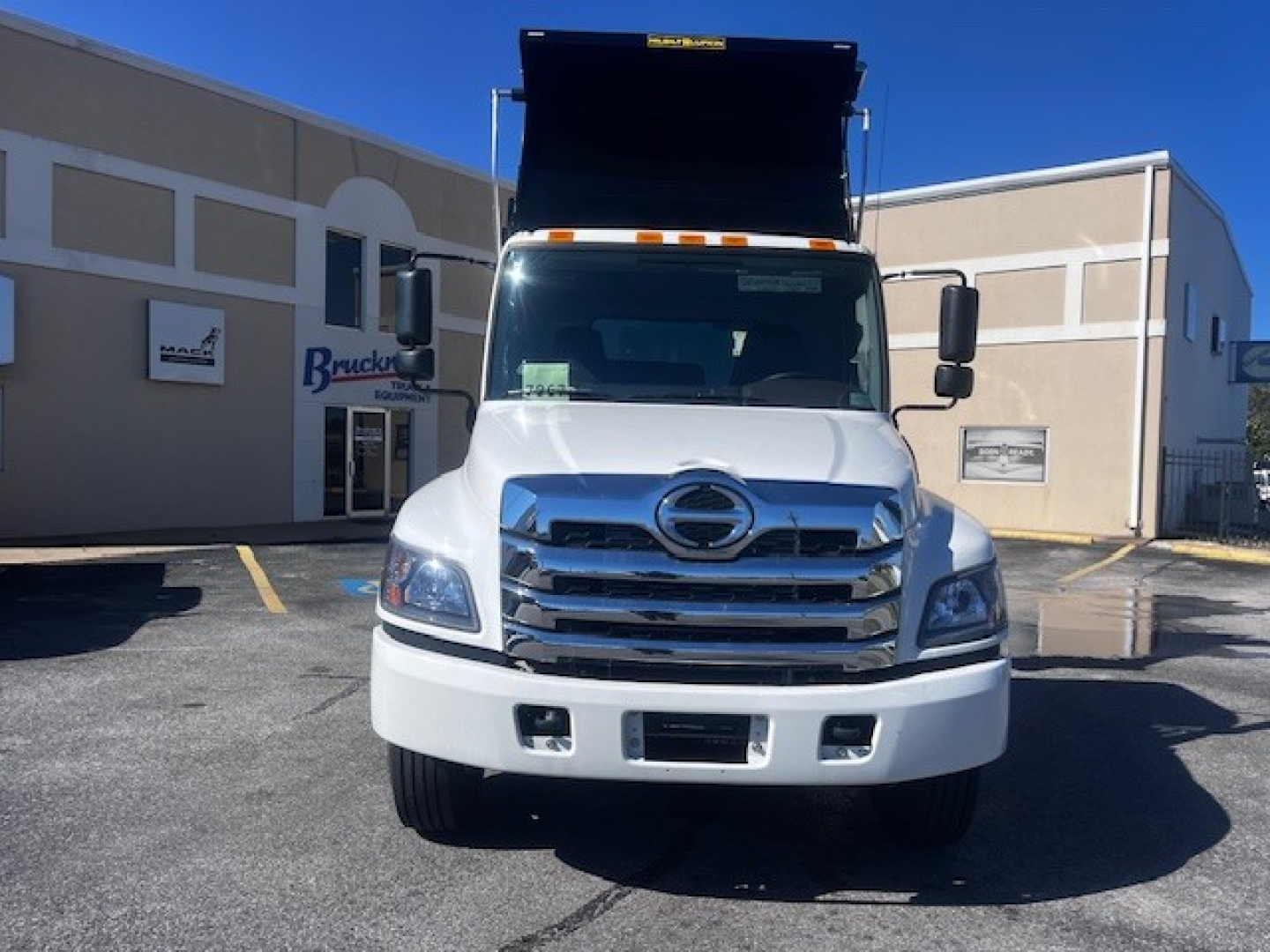 Front view of a white Hino dump truck with raised dump bed, chrome grille, and large bumper. Vehicle appears in a dealership lot.