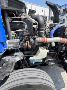 Kenworth T680 engine bay view with coolant reservoir, hoses, and intake piping visible; front wheel in foreground.