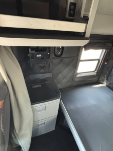 Kenworth T680 interior cab view showing sleeper area with small window, storage drawer unit, and bunk area.