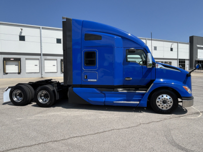Side view of a blue 2023 Kenworth T680 tractor, conventional sleeper, 6x4 configuration, parked in front of a dealership.