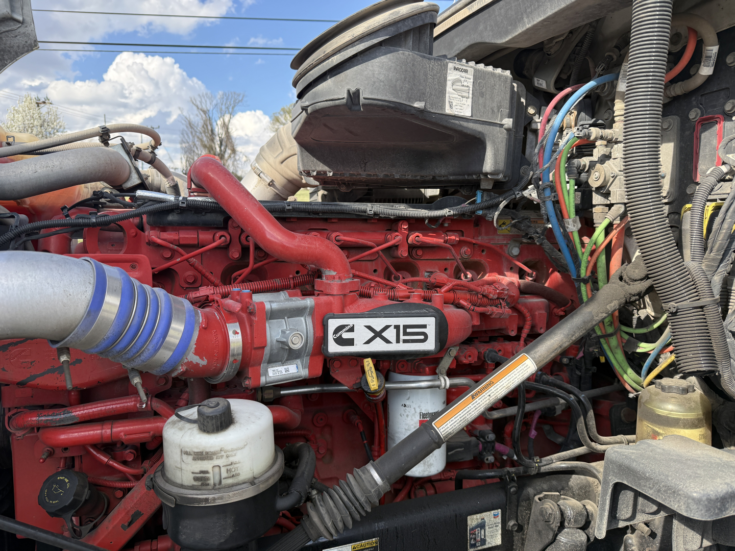 Kenworth T680 engine bay with red engine block and X15 badge, multiple hoses, wiring harnesses, and air intake piping visible.