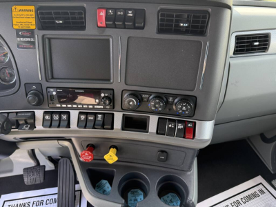 Kenworth T680 truck interior dashboard showing infotainment unit, climate controls, switches, and cup holders. Visible labels include warning sticker and various colored push buttons.