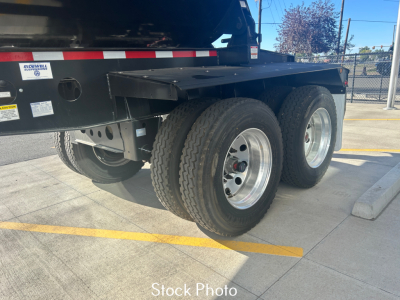 Truck trailer rear wheel area showing dual axles and black steel undercarriage with silver rims and black fenders; part of a dump trailer with stock stickers.