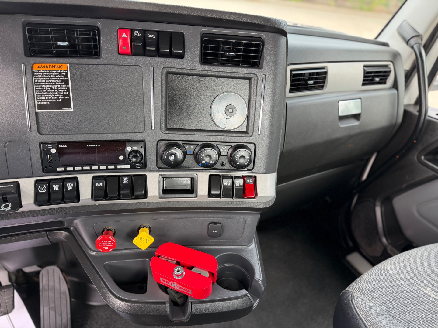 Cab interior of a 2024 Kenworth T880 showing dashboard controls, vents, switchgear, ignition, and steering column; visible yellow and red handles and a red locking device near lower dash.