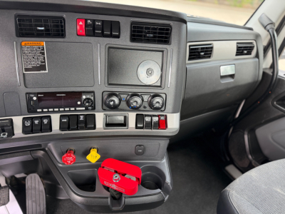 Cab interior of a 2024 Kenworth T880 showing dashboard controls, vents, switchgear, ignition, and steering column; visible yellow and red handles and a red locking device near lower dash.