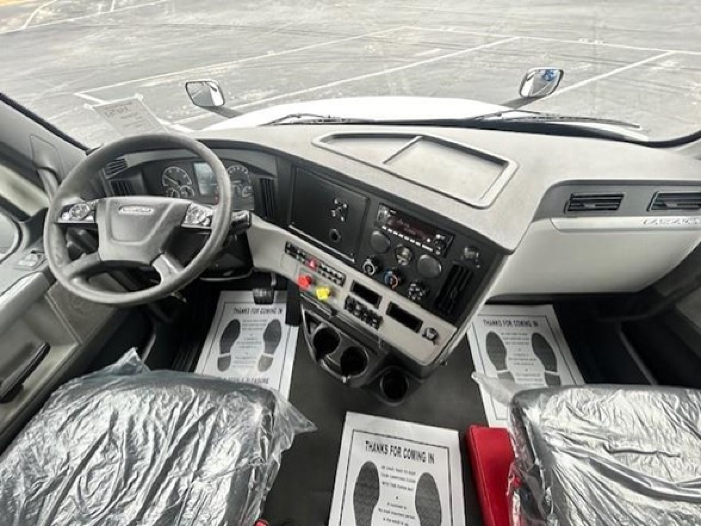 Dashboard and center console of a Freightliner Cascadia 126 truck cab showing steering wheel, gauges, controls, and two plastic-covered seats.