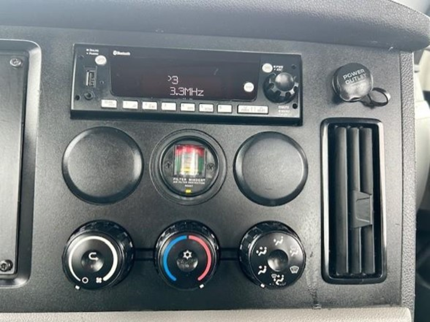 Dashboard of a Freightliner Cascadia day cab showing radio display with '3.3MHz' and control knobs, auxiliary gauges, circular vents, and a power outlet on the right.