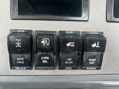 Dashboard controls showing lock, slip/air slide, lower, intr ax­le, K-PIN rel, air slide, suspend height switches in a Freightliner Cascadia dash panel.