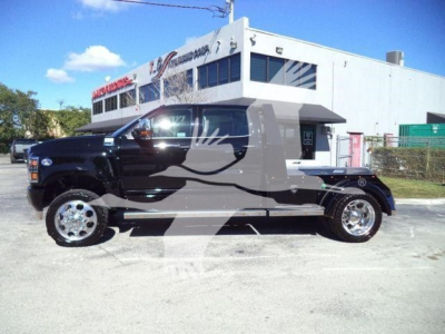 Truck with black cabin and gray flatbed, large chrome wheels, branding on side, parked in lot in front of a white building.