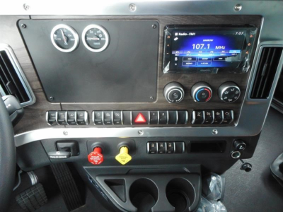 Dashboard of a Western Star 47X showing gauges, touchscreen infotainment displaying AM 107.1, climate controls, and various switches; red and yellow hazard/warning buttons visible.