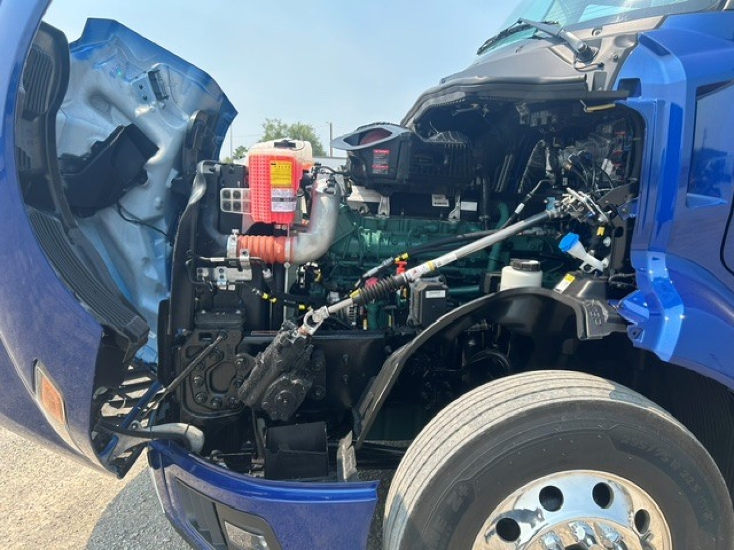 Volvo VNL64T840 engine bay showing open hood with diesel engine and various components including air intake, hoses, and battery; blue tractor unit exterior.