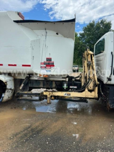 Truck cab and dump body showing rear hitch and hydraulic arm for garbage truck, side view with warning labels on hitch area.